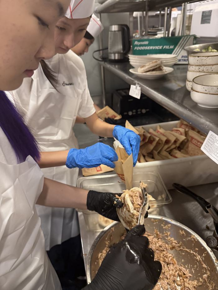 Volunteers in chef hats filling paper bags with hot food in a commercial kitchen.