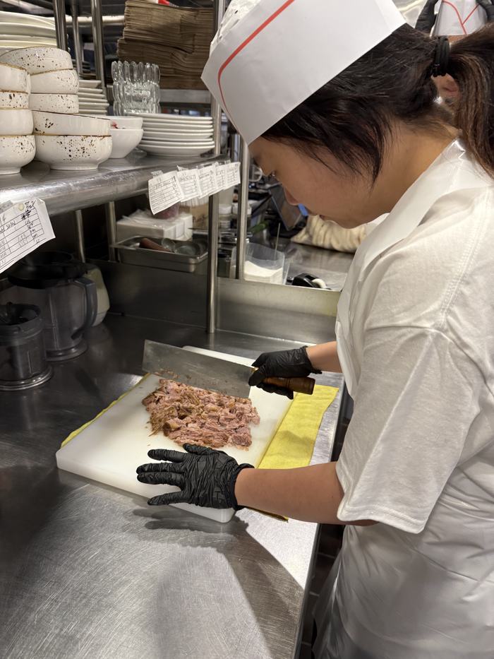 A volunteer in a chef hat chopping cooked meat on a board in the kitchen.
